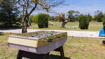 Una mesa de fútbolín en un entorno exterior con árboles y un parque infantil al fondo.