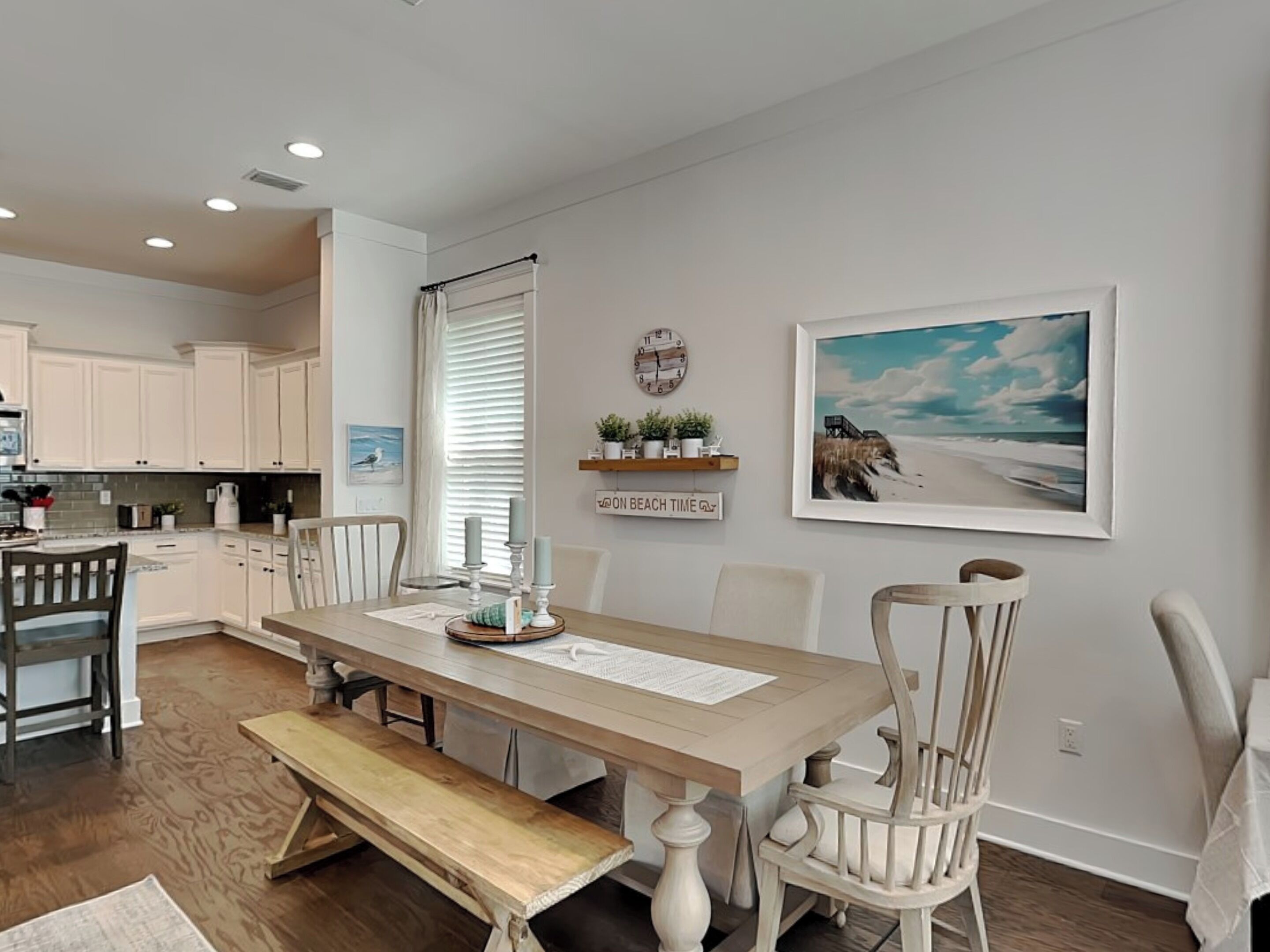 Una cocina con armarios blancos, una mesa de comedor de madera y un banco. Hay un reloj en la pared y un cuadro con una escena de playa.