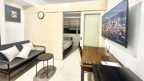 Ein gemütlicher Wohnraum mit Sofa, kleinem runden Tisch, Esstisch aus Holz, Wand-Fernseher und einem Bett mit Fenster.