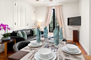 Ein Essbereich mit einem Tisch für vier Personen, weißen Tellern, Gläsern und Besteck. Es gibt ein schwarzes Ledersofa mit Türkis-Kissen, einen kleinen Beistelltisch mit einer Pflanze und einen an der Wand montierten Fernseher.