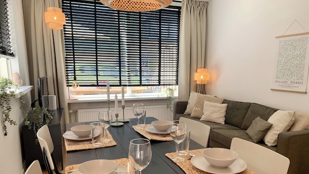 A dining area with a dark wooden table set for four, surrounded by a sofa and a window with blinds.