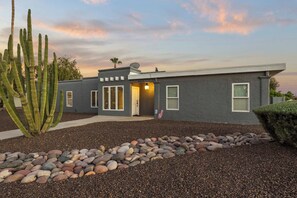 A modern house with a grey exterior, a well-maintained garden with a cactus, and a pebble pathway leading to the entrance.