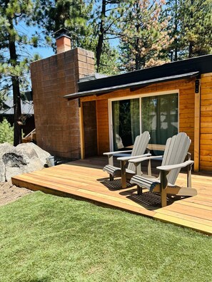 Eine Holzterrasse mit Adirondack-Stühlen, einem Ziegelkamin und einer Glastür, die zu einem überdachten Bereich führt.