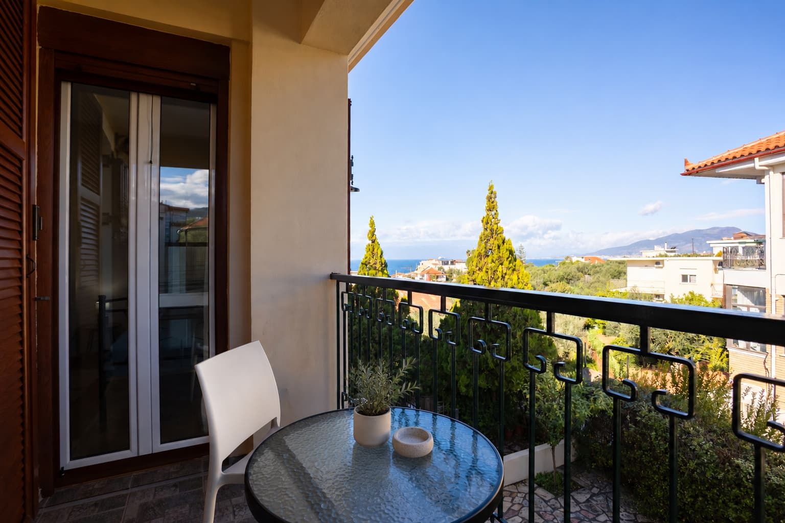 A balcony with a glass door, a round table with a potted plant, and a view of the sea and buildings.