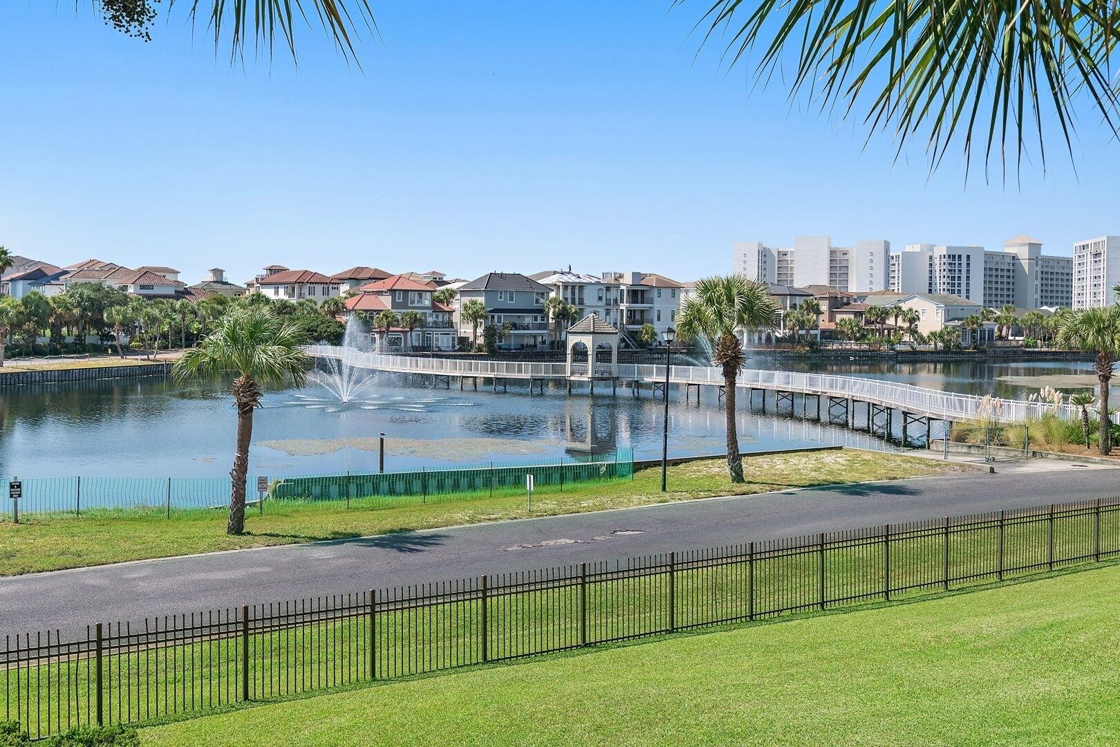 Un parque frente al agua, con una fuente, palmeras, un puente peatonal y edificios residenciales al fondo.