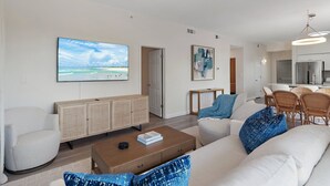 A modern living room with a large flat-screen TV, a rattan console, a coffee table, and a dining area with wicker chairs. - Terrace At Pelican Beach 0107 3 Bedroom Condo (Destin)