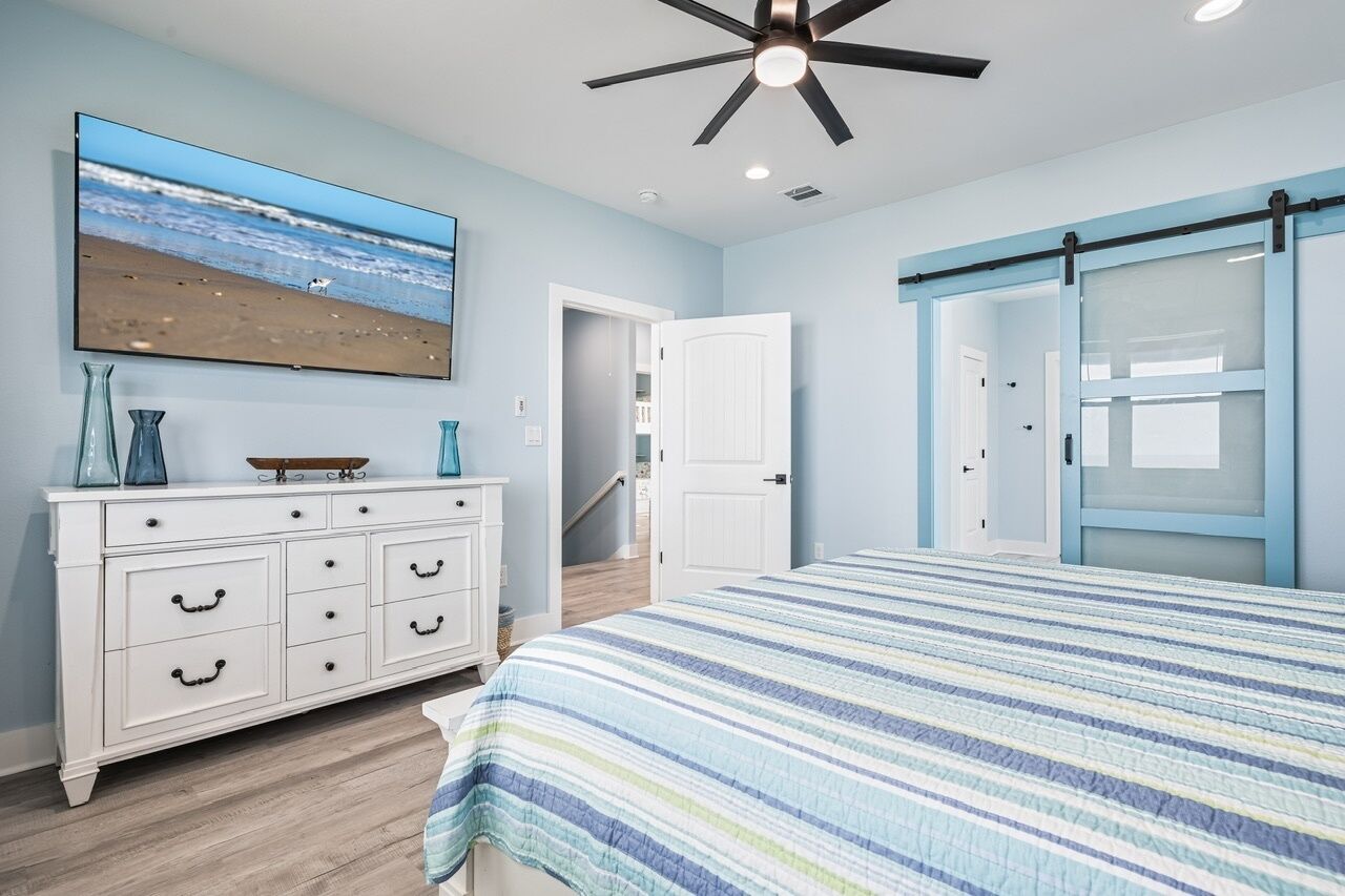 A bedroom with a bed, a dresser, a TV, a ceiling fan, and a door leading to another room.