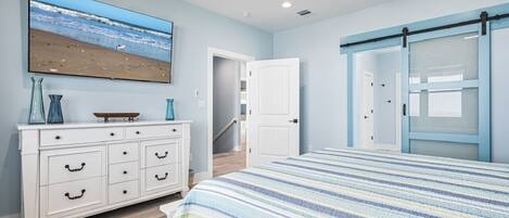 A bedroom with a bed, a dresser, a TV, a ceiling fan, and a door leading to another room.