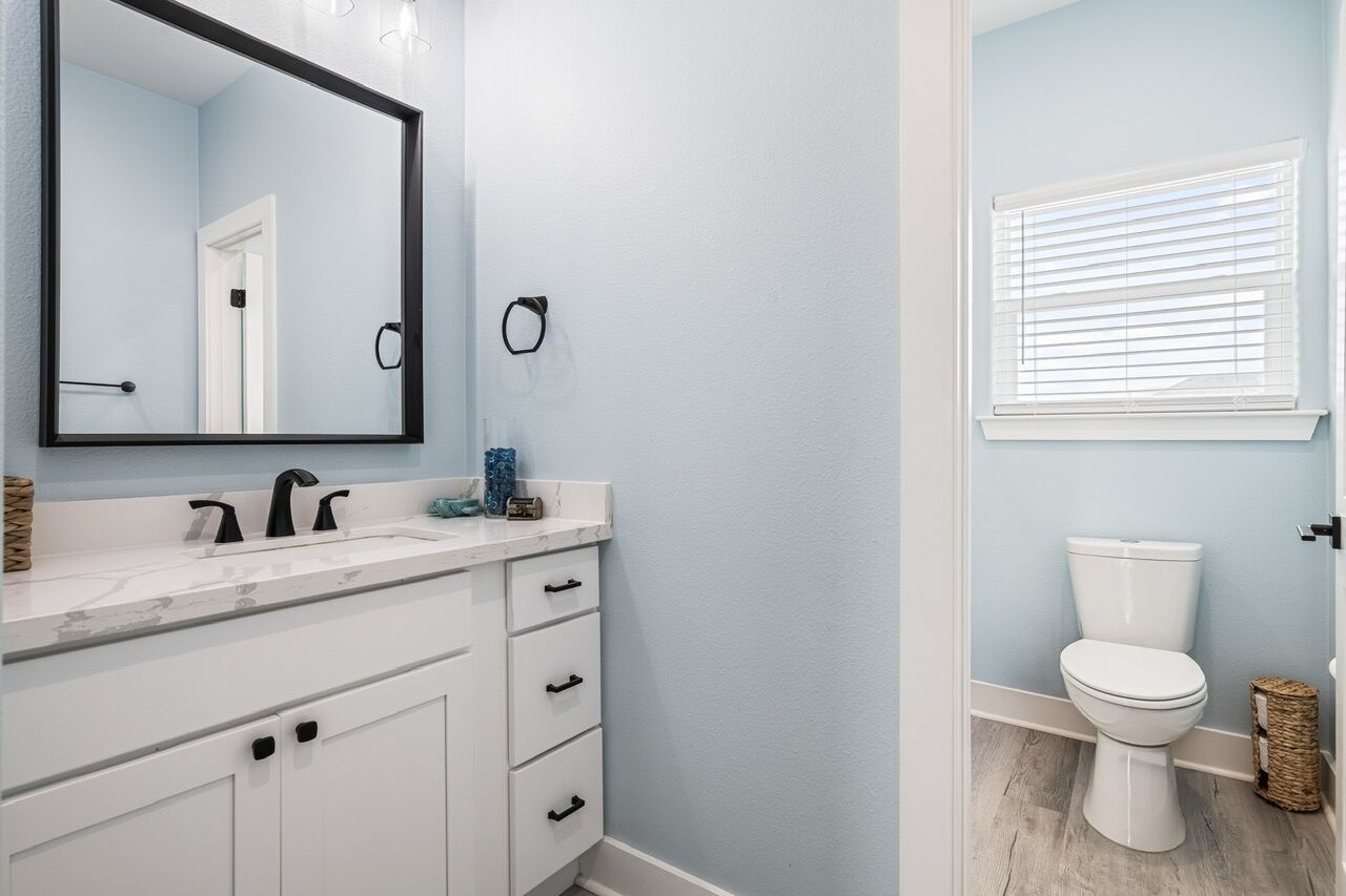 A bathroom with a white vanity, a mirror, a toilet, and a window with blinds.