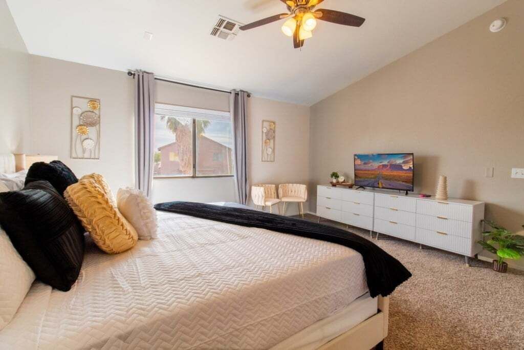 Een slaapkamer met een bed, een plafondventilator, een televisie, een dressoir en een raam met gordijnen.