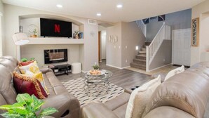 A modern living room with a leather sofa, a glass coffee table, and a flat-screen TV mounted above a fireplace. - Casa Aria 4br Home & Heated Pool 4 Bedroom Home (Casa Grande)