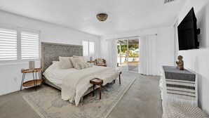 Un dormitorio con una cama grande, una mesita de noche pequeña, un banco, una alfombra, un aparador y una ventana con persianas.