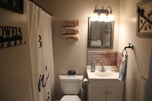 A bathroom with a toilet, sink, and mirror. There are towels hanging on the wall and a shower curtain with a number. - Step Back In Time to a charming 2-bedroom lower level house in adorable Eudora (Eudora)