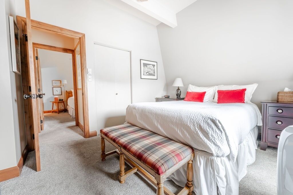 Une chambre à coucher comprenant un lit, un banc, une table de chevet, une lampe et une porte donnant sur une autre pièce.