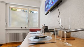 A tidy dining area with a wooden table, a television mounted on the wall, and a window with blinds. - Crans-Montana - Studio ski 2 pers - 3 min Cry d'Er (Crans-Montana)