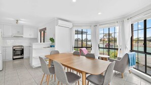 A modern kitchen with white cabinets, a built-in oven, and a dining area with a wooden table and grey chairs. - 17 Matthew Flinders Dve Encounter Bay (Encounter Bay)