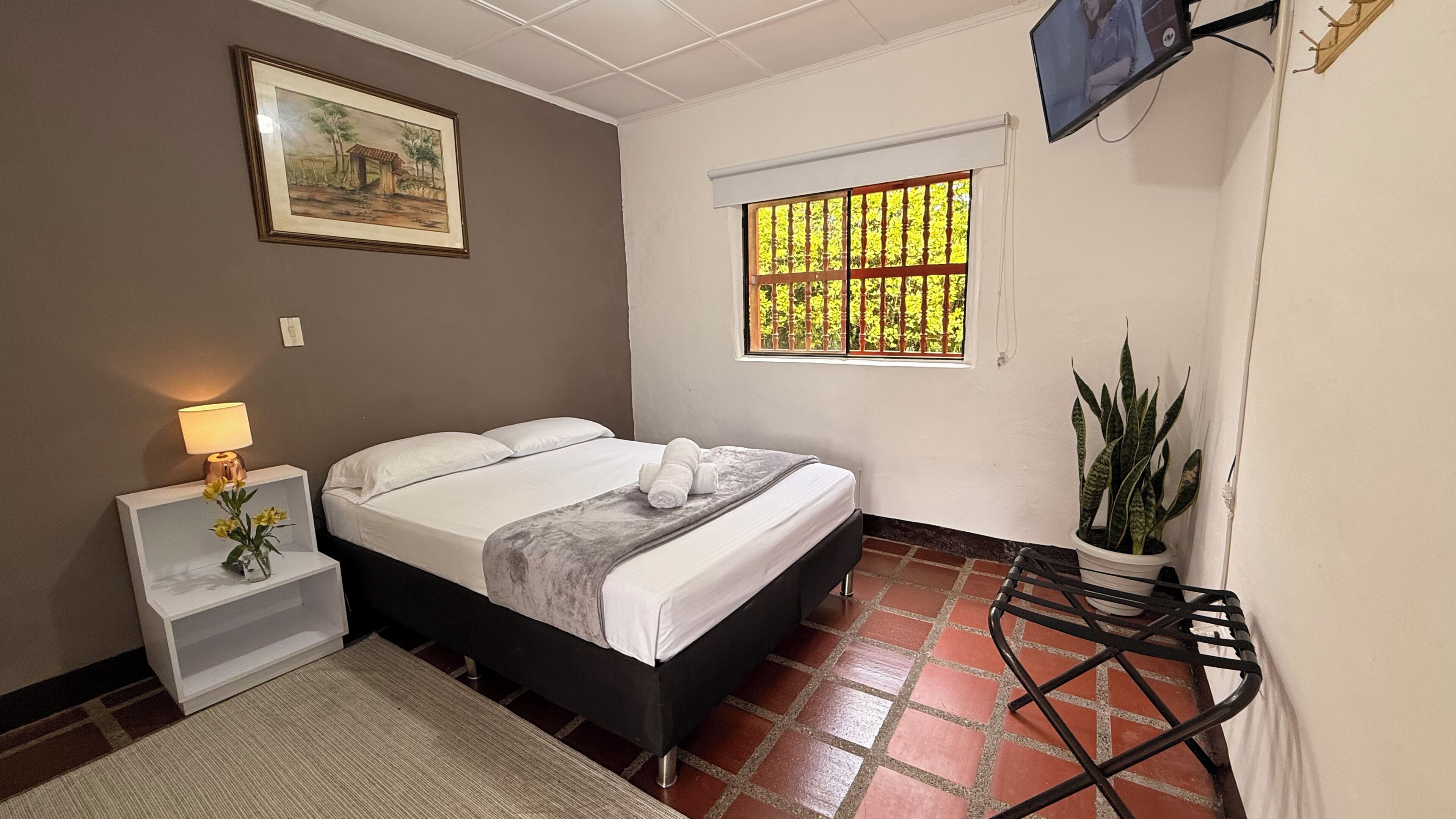 A bedroom with a bed, a nightstand, a lamp, a window with bars, a television, and a potted plant.