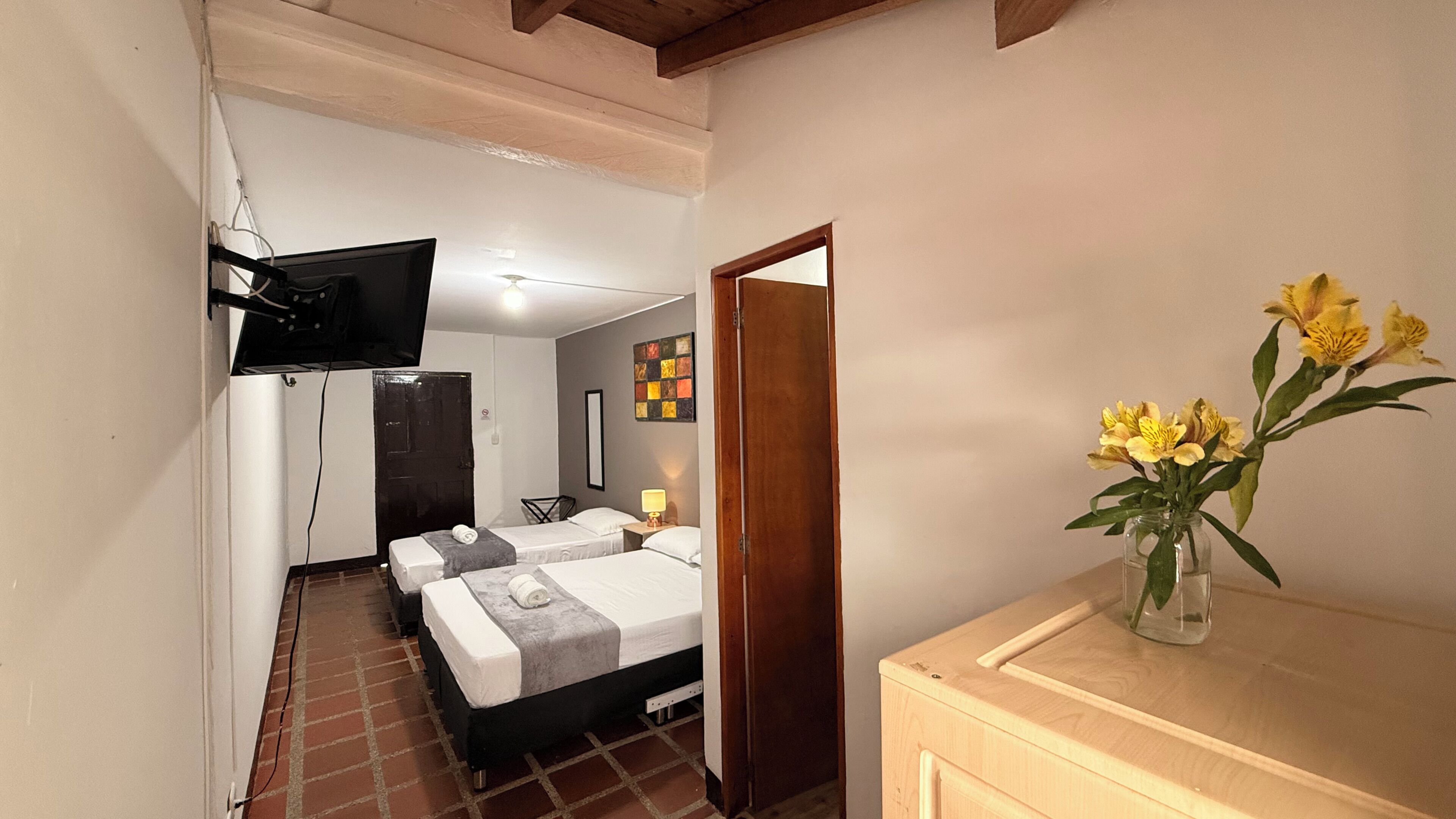 A hotel room with a bed, a TV mounted on the wall, a small table with a lamp, and a vase with flowers on a dresser.