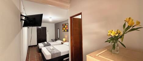 A hotel room with a bed, a TV mounted on the wall, a small table with a lamp, and a vase with flowers on a dresser.