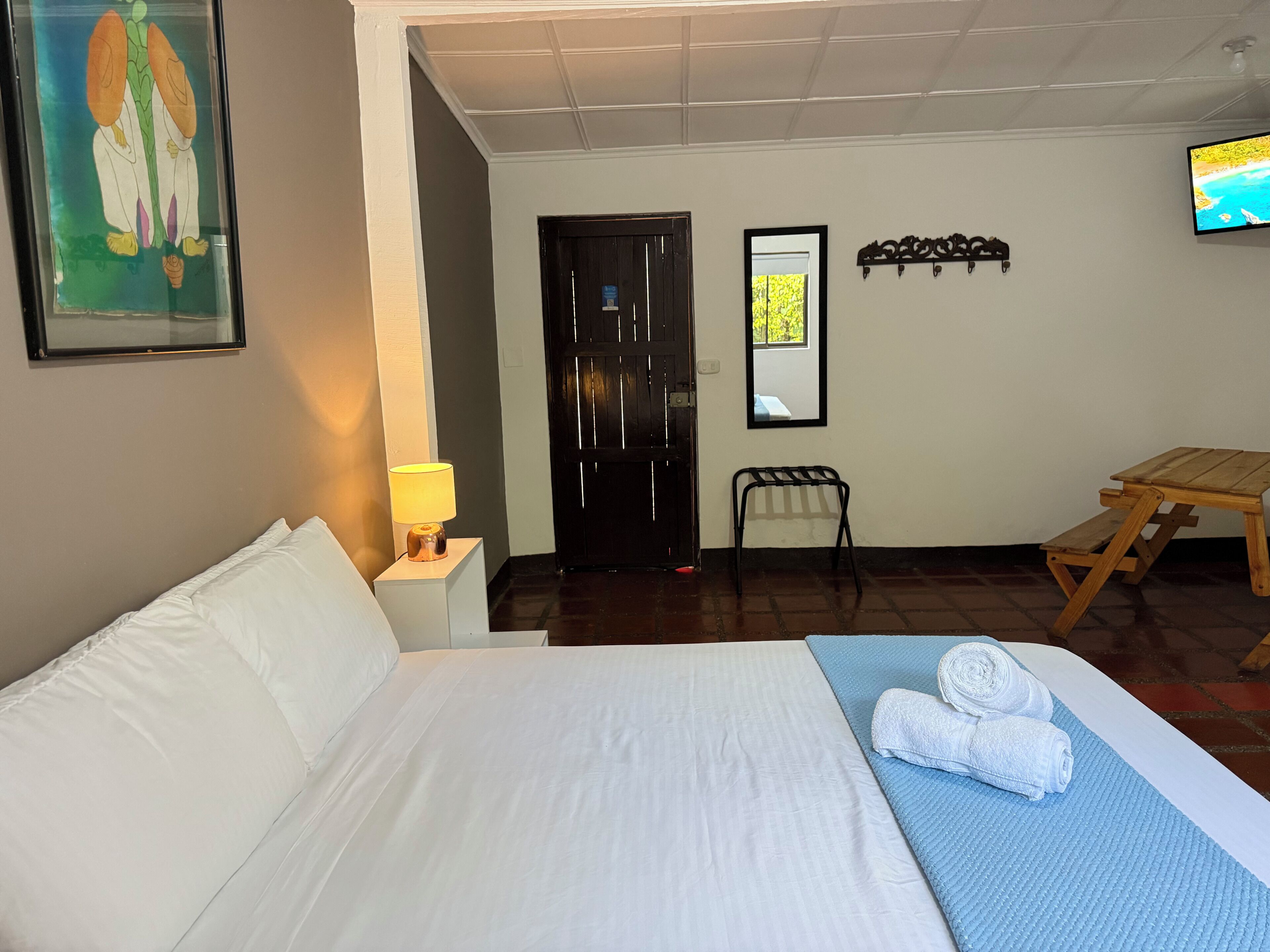 Une chambre d’hôtel avec un lit, une table de chevet avec une lampe, une table en bois, une chaise, un miroir et un tableau accroché au mur.