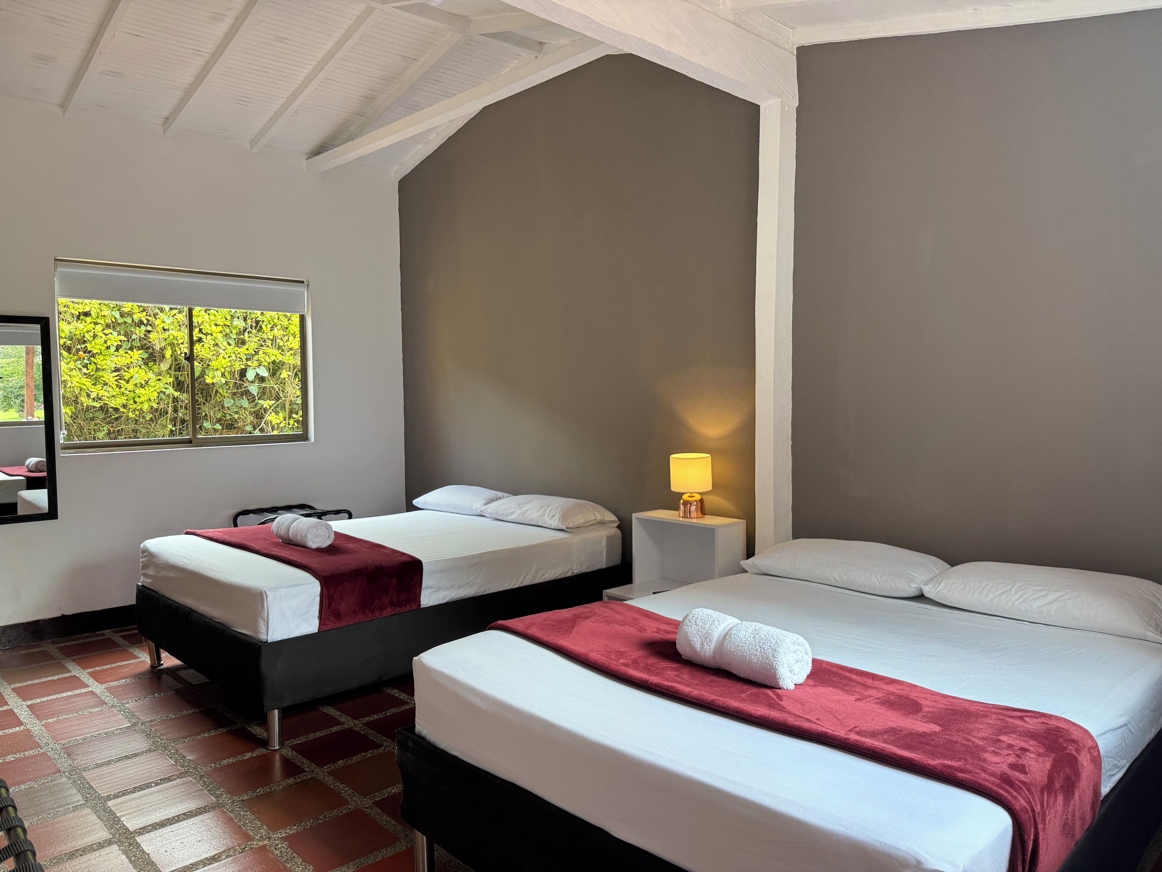 Een hotelkamer met twee bedden, een raam met uitzicht op groen, een nachtkastje met een lamp en een vloer met een bakstenen patroon.