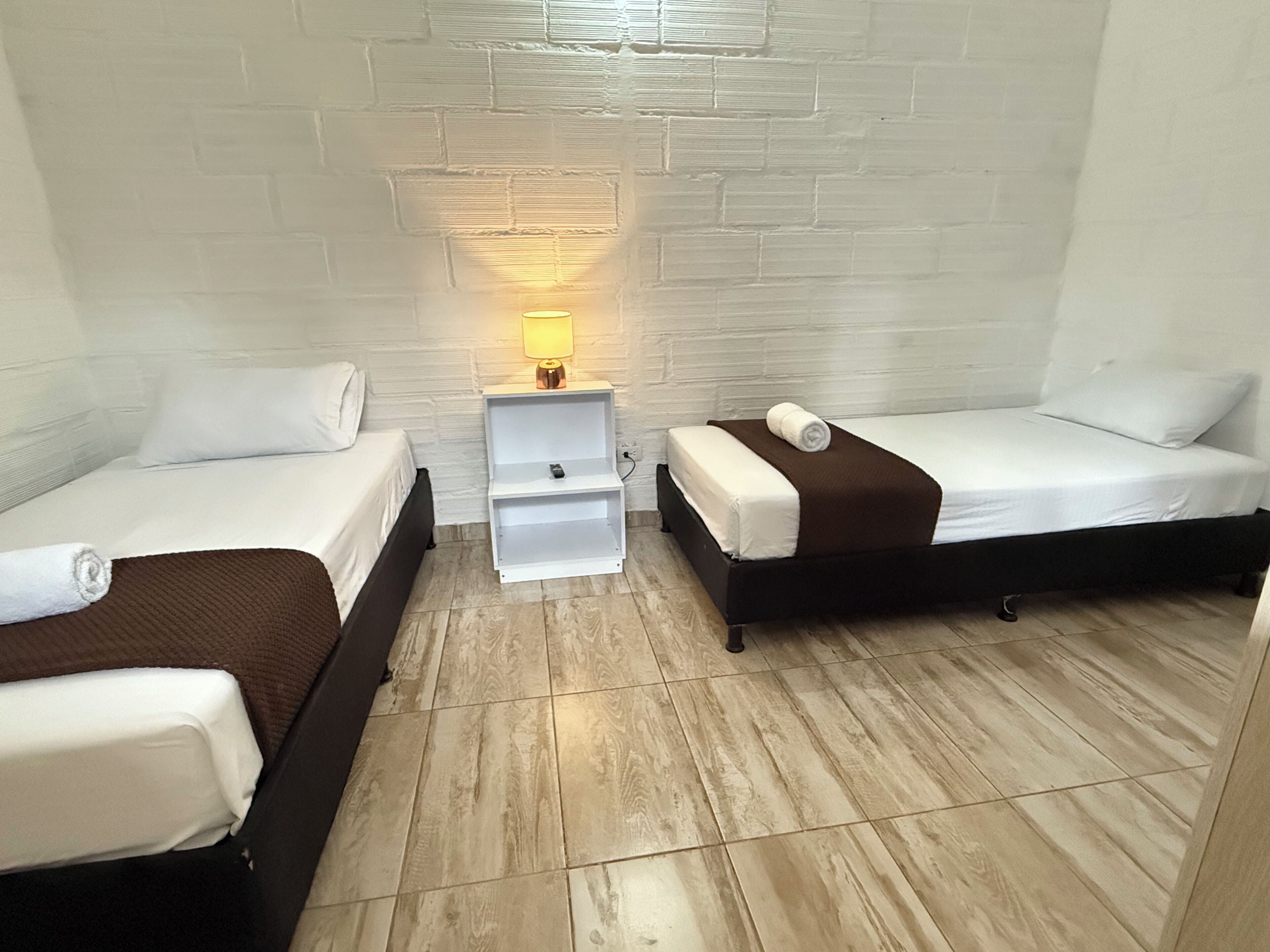 A small, clean room with two single beds, a nightstand with a lamp, and a white brick wall.