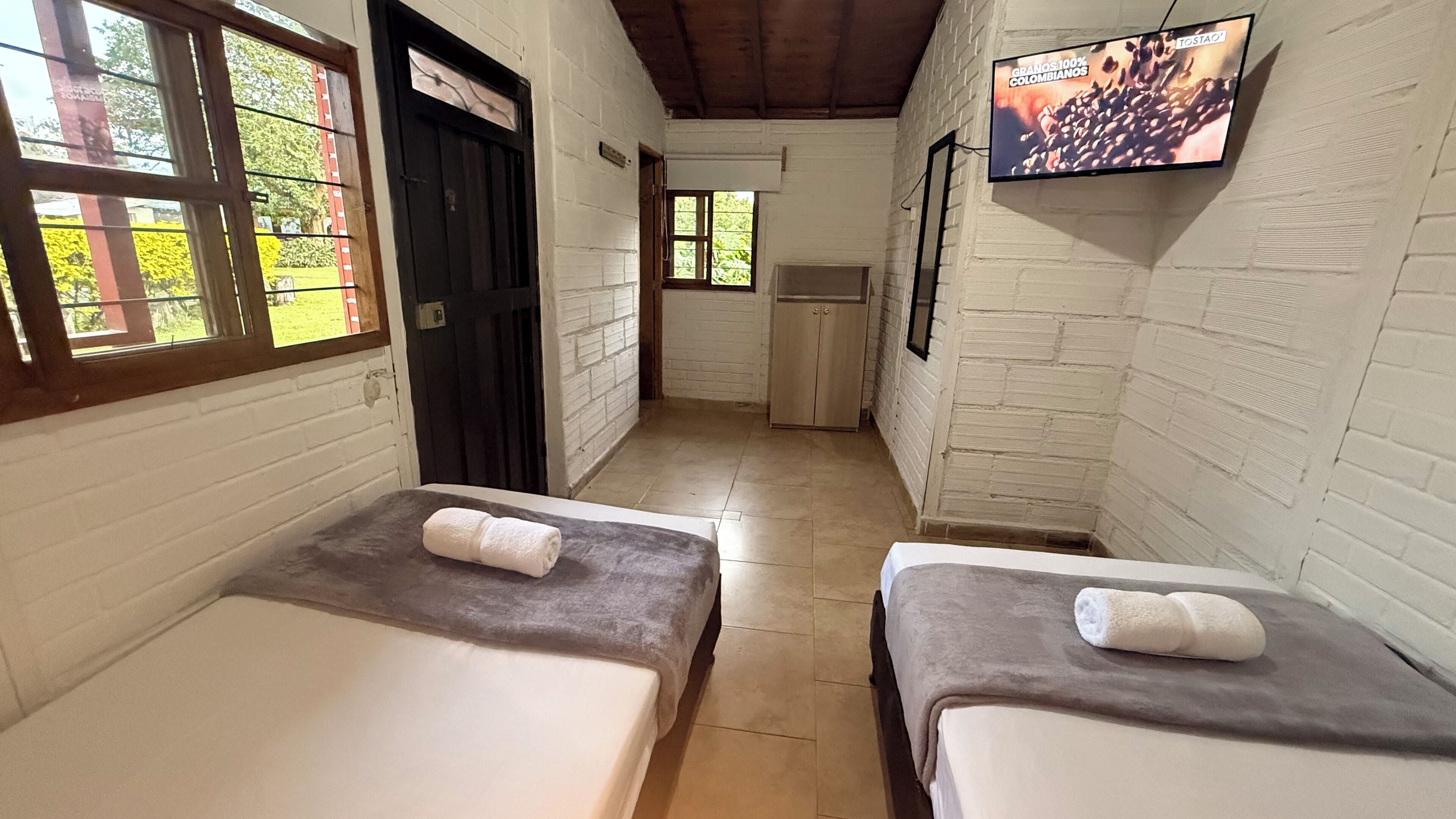 A room with two beds, a TV on the wall, and a window with a view of greenery.