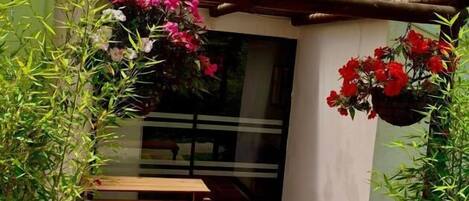 A small patio with a tiled floor, a wooden table, and chairs, surrounded by plants and a hanging flower basket.