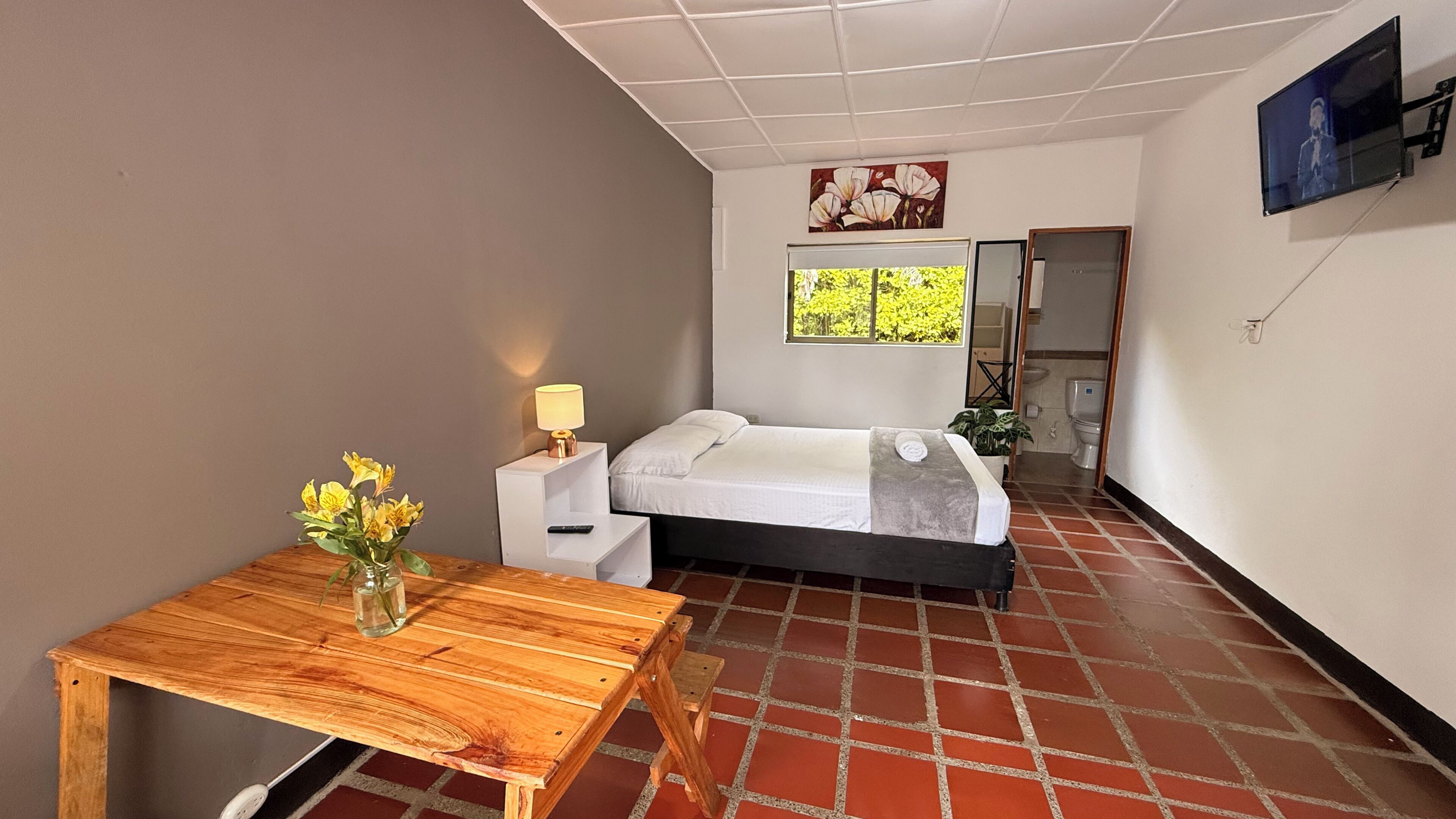 Una habitació amb un llit, una taula petita amb un ram de flors, un televisor penjat a la paret i una finestra amb vista a vegetació.