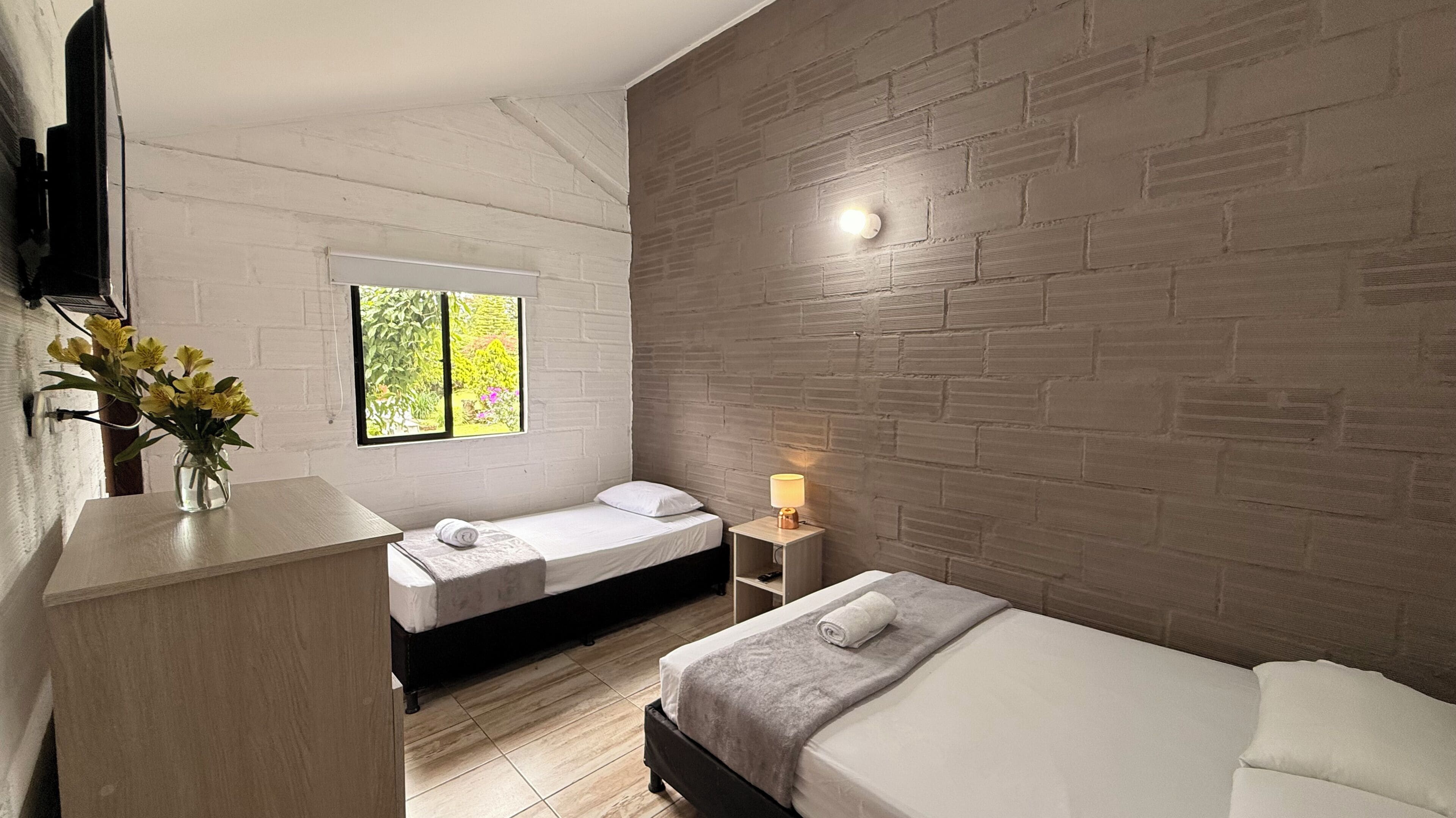 A hotel room with two beds, a window with a view of greenery, a bedside lamp, and a flat-screen TV mounted on the wall.