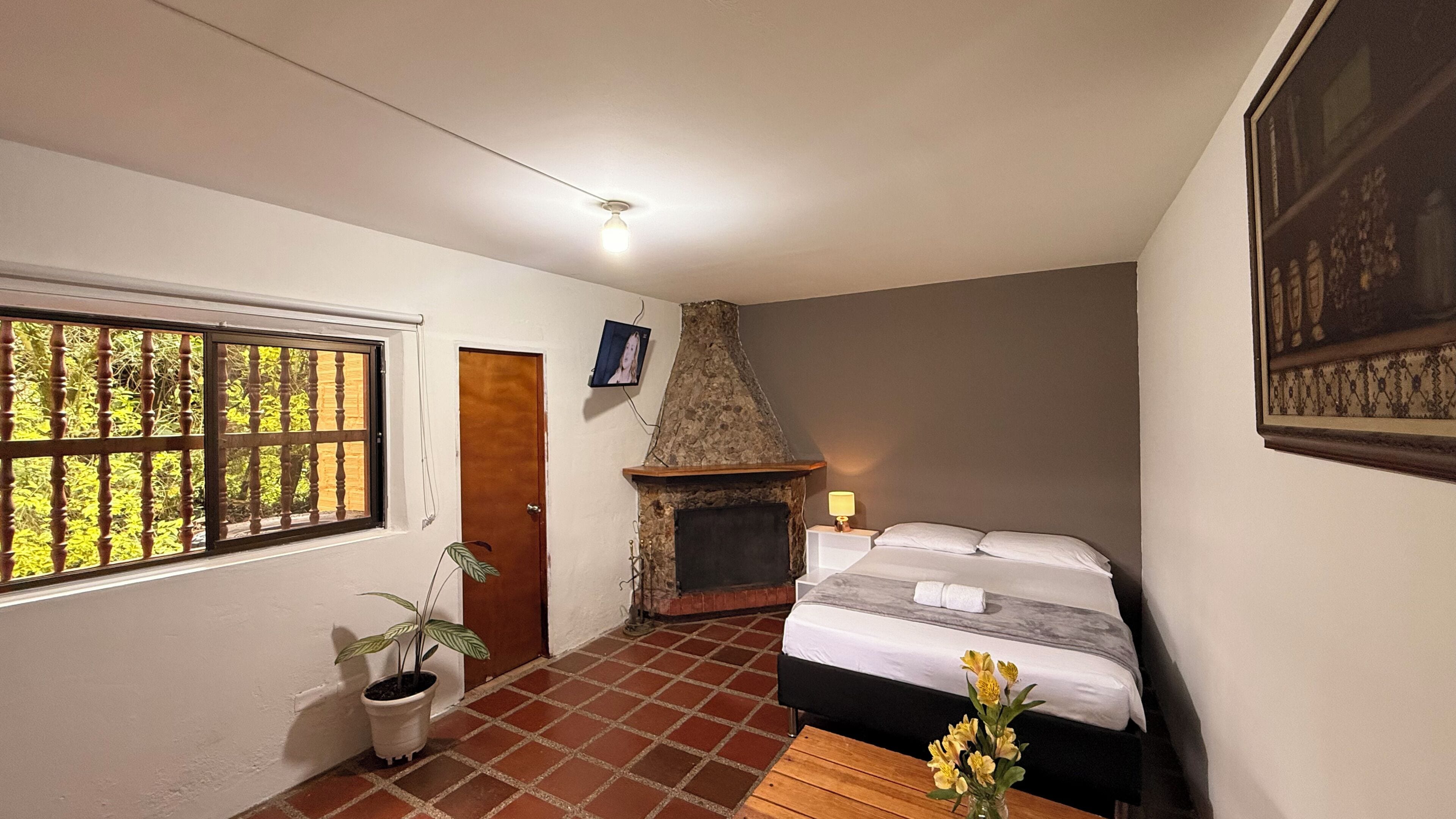 A bedroom with a stone fireplace, a bed with white bedding, a small table with flowers, and a window with a view of greenery.