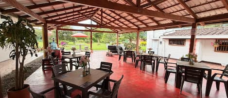 An outdoor patio area with wooden tables and chairs, surrounded by a red floor and a metal roof structure.