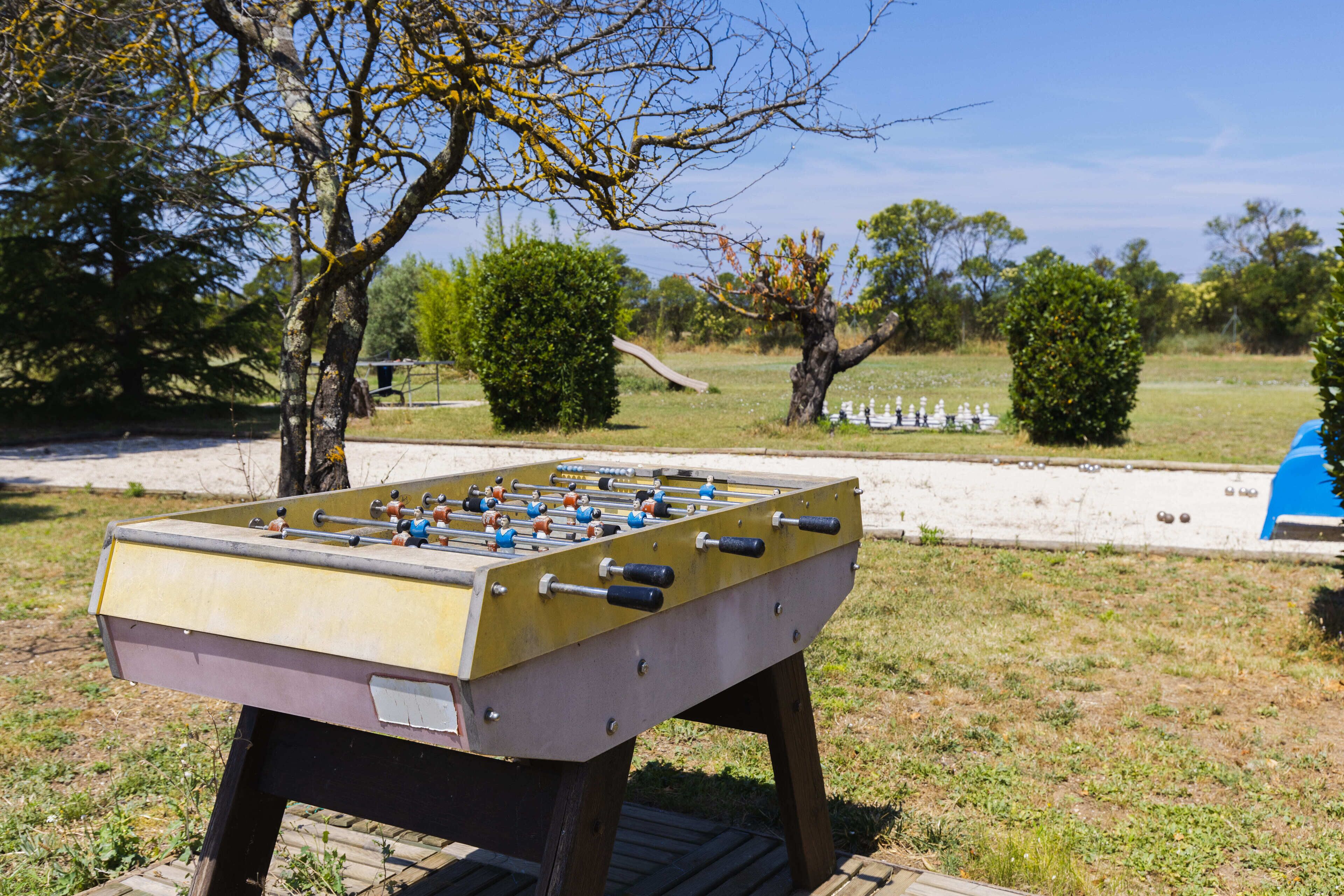 Una mesa de fútbolín en un entorno exterior con árboles y un parque infantil al fondo.