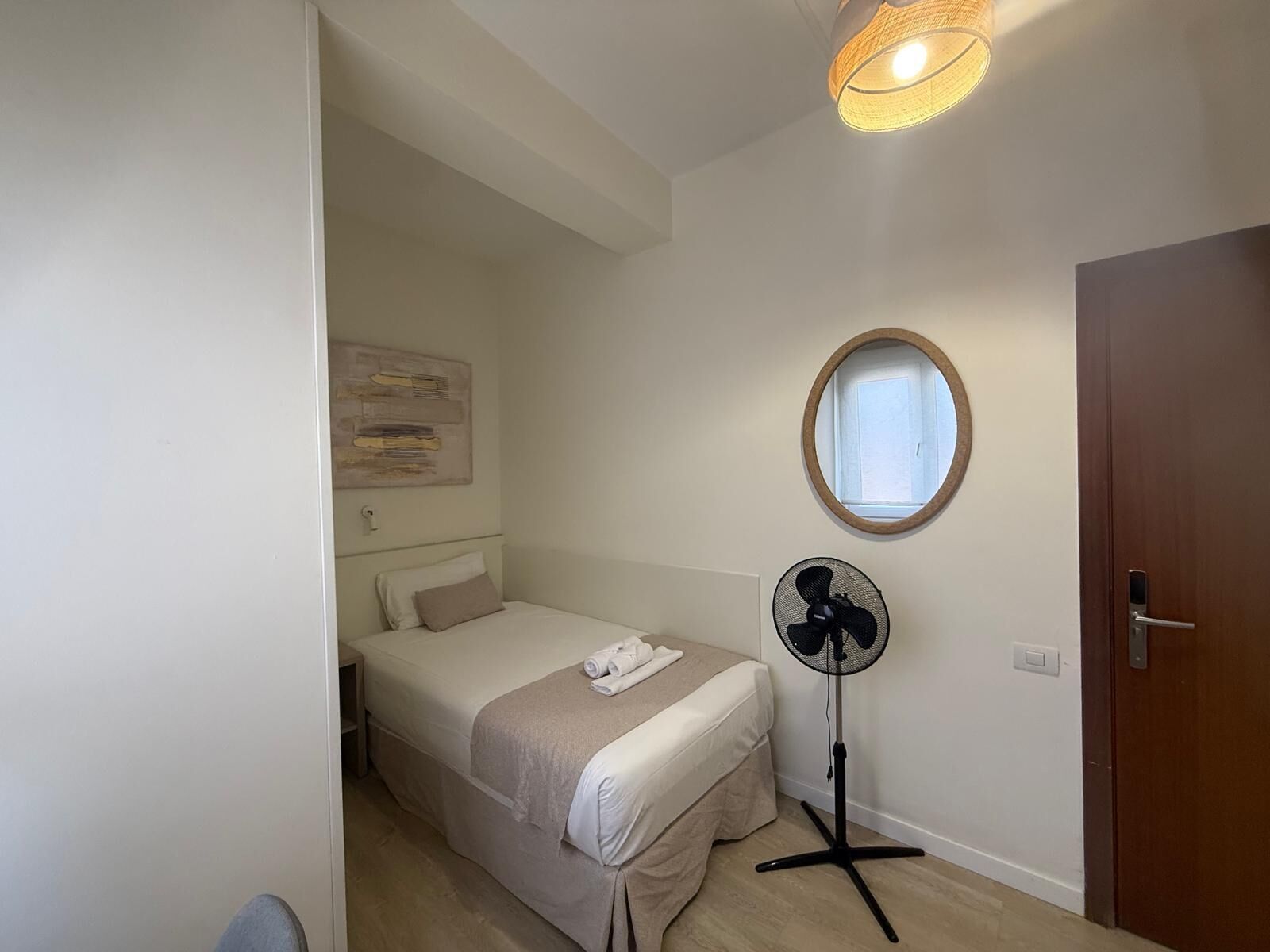 A small, neatly arranged bedroom with a bed, a standing fan, a window, and a door.