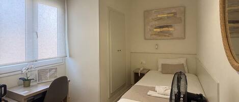 A bedroom with a bed, a desk, a chair, a standing fan, a window with blinds, and a wall-mounted mirror.