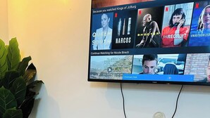 Un saló amb un televisor de pantalla plana fixat a la paret que mostra programes de Netflix, una taula de consola de fusta amb una caixa de perfum Chanel, una planta en test i una catifa amb dibuixos.