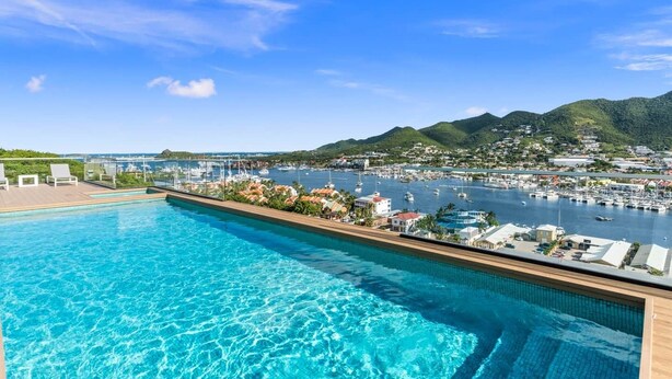 Una piscina con agua azul cristalina, rodeada por una terraza de madera y sillas de descanso, con vistas a un puerto deportivo con barcos y un paisaje montañoso.