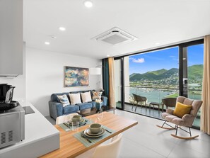Un salon moderne comprenant un coin repas, un canapé, une machine à café et une vue sur un port avec des bateaux et des montagnes.