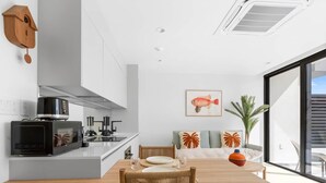 A modern kitchen with a microwave, toaster, and kettle. There is a dining area with a wooden table and chairs. A fish illustration is on the wall, and a wooden birdhouse is mounted on the wall.