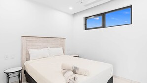 A modern bedroom with a large bed, a small side table, and a window with a clear view of the sky.