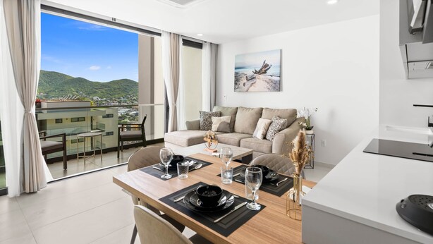 A modern living room with a dining area, large windows, and a view of a mountain. - D414 – 1-Bedroom Apartment with Balcony (Sint Maarten)