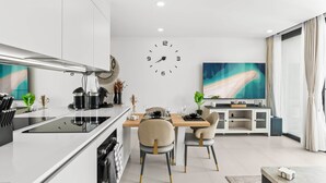 A modern kitchen with a central island, a dining area with two chairs, a large wall clock, and a flat-screen TV. - D414 – 1-Bedroom Apartment with Balcony (Sint Maarten)