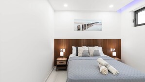 A modern bedroom with a large bed, wooden headboard, two bedside tables with lamps, a wall-mounted painting, and a window with blinds. - D414 – 1-Bedroom Apartment with Balcony (Sint Maarten)