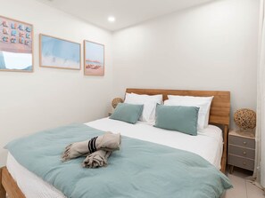 A bedroom with a wooden bed, white bedding, and a teal throw blanket. There are two framed pictures on the wall and a small wicker side table.