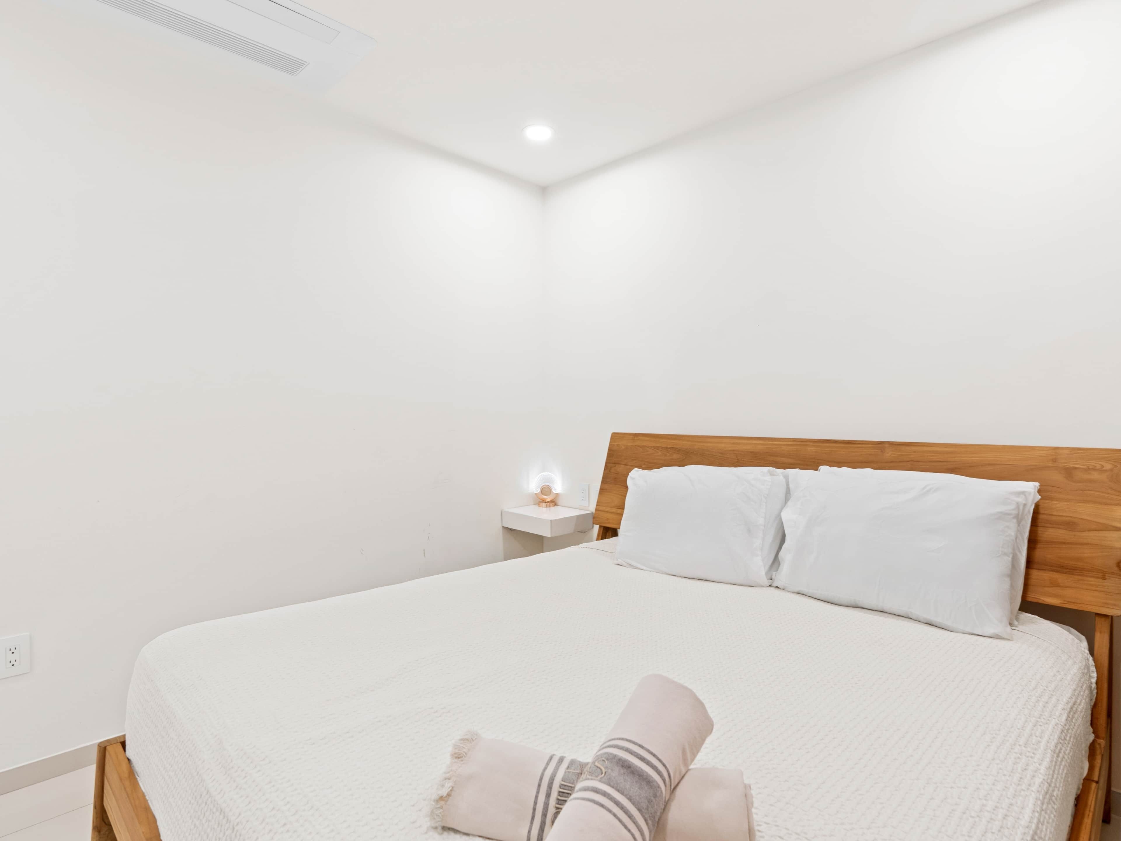 A neatly made bed with white bedding and a wooden headboard in a bright, minimalistic room.