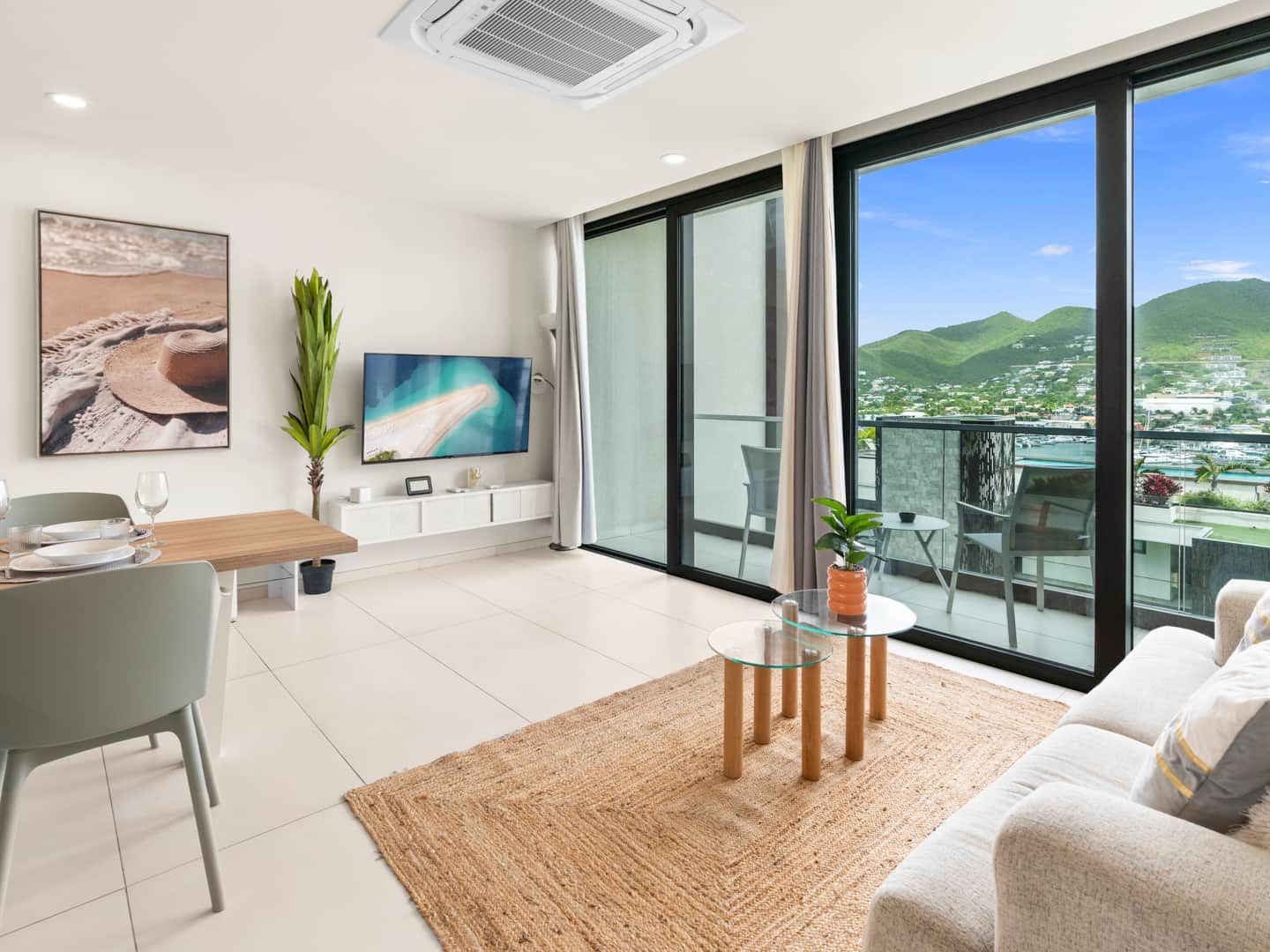 A modern living room with a flat-screen TV, a dining area with a table and chairs, a large window with a view of a coastal landscape, and a balcony with a glass table and chairs.