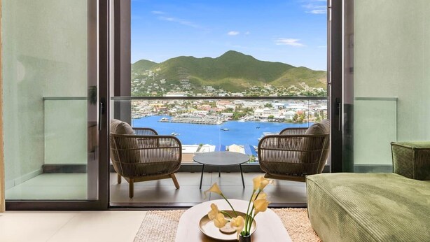 Un balcon avec vue sur une ville côtière et les montagnes. On y trouve deux fauteuils en osier et une petite table sur laquelle est posé un vase de fleurs.