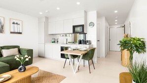Une cuisine moderne avec des meubles blancs, un four à micro-ondes et une machine à café. On y trouve un coin repas avec une table en bois et des chaises vertes. Un canapé vert et une table basse se trouvent dans l’espace salon.