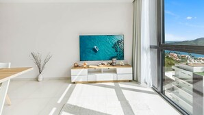 A modern living room with a large window, a flat-screen TV, a wooden dining table, and a minimalist white console. - C333 - Charming Apartment with Lagoon View (Sint Maarten)