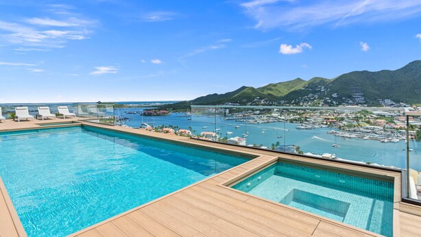 A pool area with clear blue water, wooden deck, and a view of a marina with boats and a hilly landscape in the background.